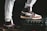 A pair of stylish high-top sneakers with brown, black, and white color combinations on different surfaces stands prominently. One shoe rests on a sturdy black case while the person is wearing light gray sweatpants.