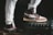 A pair of stylish high-top sneakers with brown, black, and white color combinations on different surfaces stands prominently. One shoe rests on a sturdy black case while the person is wearing light gray sweatpants.
