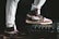 A pair of stylish high-top sneakers with brown, black, and white color combinations on different surfaces stands prominently. One shoe rests on a sturdy black case while the person is wearing light gray sweatpants.