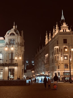 Illuminated European architecture at night, with ornate buildings featuring domes and spires. Warm street lights cast a glow over the cobblestone streets. In the foreground, people are walking and one person is pushing a stroller.