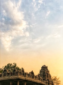A beautifully crafted ancient temple with intricate carvings and sculptures on its facade. The sky above features a mix of soft clouds with warm, golden hues, suggesting a sunset or sunrise. Lush green trees surround the temple, adding to the serene atmosphere.