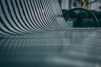 A close-up shot of a metallic bench with parallel slats, creating a repetitive pattern. The focus is shallow, highlighting the texture and lines of the bench, while the background appears blurred, suggesting an outdoor setting.