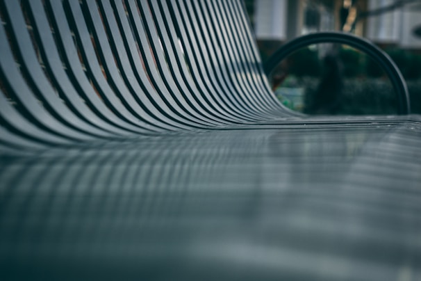 A close-up shot of a metallic bench with parallel slats, creating a repetitive pattern. The focus is shallow, highlighting the texture and lines of the bench, while the background appears blurred, suggesting an outdoor setting.