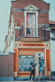 A brick building features a painted mural creating the illusion of a cafe facade. It shows a woman standing on a balcony holding a book. Below, two people adjust the mural that includes painted shelves of pastries and a door. One person stands on a ladder, while the other gives instructions.
