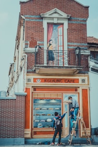 A brick building features a painted mural creating the illusion of a cafe facade. It shows a woman standing on a balcony holding a book. Below, two people adjust the mural that includes painted shelves of pastries and a door. One person stands on a ladder, while the other gives instructions.
