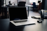A laptop with a blank screen is positioned centrally on a dark desk, with a phone and a computer mouse nearby. The background is a blurred office setting with some indistinct furniture and lighting.
