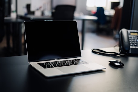 A laptop with a blank screen is positioned centrally on a dark desk, with a phone and a computer mouse nearby. The background is a blurred office setting with some indistinct furniture and lighting.