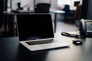 A laptop with a blank screen is positioned centrally on a dark desk, with a phone and a computer mouse nearby. The background is a blurred office setting with some indistinct furniture and lighting.