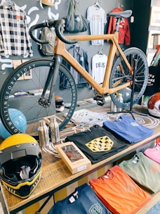 A wooden bicycle with black tires is displayed prominently on a table. Surrounding the bicycle are various merchandise items including colorful t-shirts featuring the Deus logo, a black and yellow helmet, two stainless steel water bottles, and some clothing on a wall-mounted rack. The wall behind the table is decorated with different jackets and shirts.
