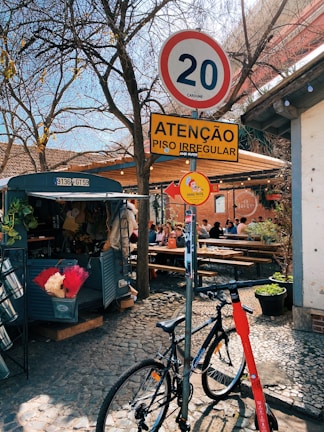 A quaint outdoor cafe scene features a cobblestone pathway with a parked bicycle and scooter by a post displaying various signs, including speed limit and caution warnings. Nearby, people enjoy their time sitting at wooden picnic-style tables under a canopy, surrounded by trees and a food truck stocked with items.