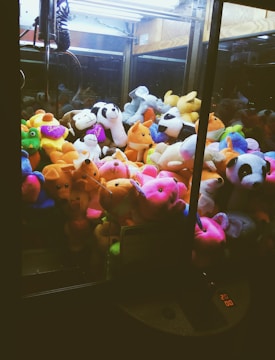 A collection of colorful stuffed animals inside a claw machine, including a panda, monkeys, foxes, and various other plush toys. The machine's interior lighting highlights the vibrant hues of the toys, creating a playful and inviting atmosphere.
