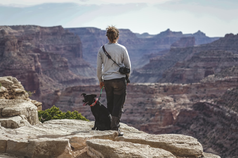 man in gray hoodie and black pants standing on rock with black labrador retriever on top