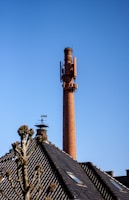 A tall brick chimney with various antennas and equipment attached, set against a clear blue sky. In the foreground, the roof of a building with black tiles is visible, along with a weather vane and a bare tree with some budding branches.