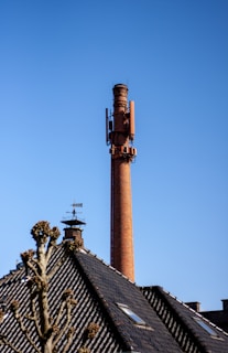 A tall brick chimney with various antennas and equipment attached, set against a clear blue sky. In the foreground, the roof of a building with black tiles is visible, along with a weather vane and a bare tree with some budding branches.