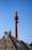 A tall brick chimney with various antennas and equipment attached, set against a clear blue sky. In the foreground, the roof of a building with black tiles is visible, along with a weather vane and a bare tree with some budding branches.