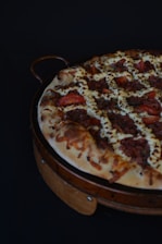 A pizza with a thick crust topped with melted cheese, slices of tomato, minced meat, and herbs, arranged in a circular wooden pizza tray.