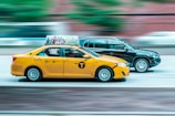 A yellow taxi and a black SUV are driving side by side on a road, with motion blur emphasizing their speed. The taxi has an advertisement on top and showcases iconic NYC taxi markings.