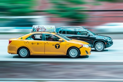 A yellow taxi and a black SUV are driving side by side on a road, with motion blur emphasizing their speed. The taxi has an advertisement on top and showcases iconic NYC taxi markings.