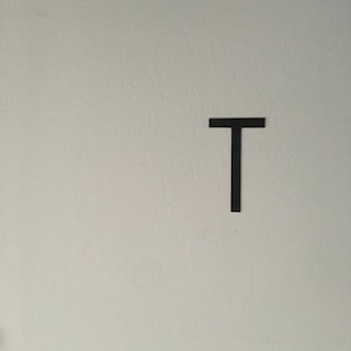 A minimalistic composition featuring a single uppercase black letter 'T' centered against a plain, light-colored wall.