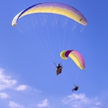 Two paragliders soaring through a clear blue sky with colorful parachutes, one in shades of purple and yellow, and the other in pink and yellow. The scene conveys a sense of freedom and adventure.