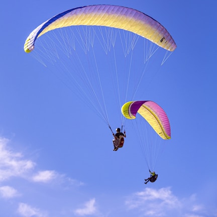 Two paragliders soaring through a clear blue sky with colorful parachutes, one in shades of purple and yellow, and the other in pink and yellow. The scene conveys a sense of freedom and adventure.