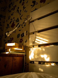 Sunlit cozy bedroom corner with vintage floral wallpaper and a distressed wooden nightstand topped with an antique lamp.