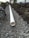 A trench filled with gravel runs parallel to a white PVC pipe, partially buried and surrounded by soil and dirt mounds on both sides. The setting appears to be outdoors near a building with trees in the background.