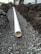 A trench filled with gravel runs parallel to a white PVC pipe, partially buried and surrounded by soil and dirt mounds on both sides. The setting appears to be outdoors near a building with trees in the background.