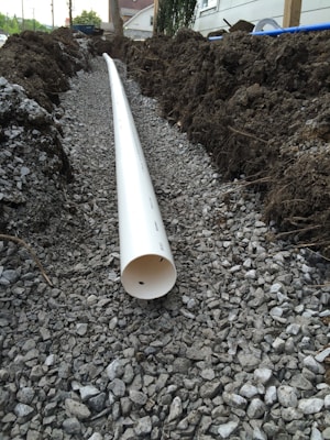 A trench filled with gravel runs parallel to a white PVC pipe, partially buried and surrounded by soil and dirt mounds on both sides. The setting appears to be outdoors near a building with trees in the background.