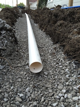 A trench filled with gravel runs parallel to a white PVC pipe, partially buried and surrounded by soil and dirt mounds on both sides. The setting appears to be outdoors near a building with trees in the background.