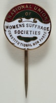 A vintage pin with text that reads 'National Union of Women's Suffrage Societies, Constitutional Non Party'. The pin has a circular design with a metallic needle attached for wearing. The text is arranged within colored segments at the edges.
