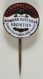 A vintage pin with text that reads 'National Union of Women's Suffrage Societies, Constitutional Non Party'. The pin has a circular design with a metallic needle attached for wearing. The text is arranged within colored segments at the edges.