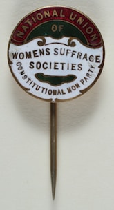A vintage pin with text that reads 'National Union of Women's Suffrage Societies, Constitutional Non Party'. The pin has a circular design with a metallic needle attached for wearing. The text is arranged within colored segments at the edges.