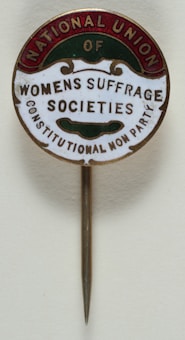 A vintage pin with text that reads 'National Union of Women's Suffrage Societies, Constitutional Non Party'. The pin has a circular design with a metallic needle attached for wearing. The text is arranged within colored segments at the edges.