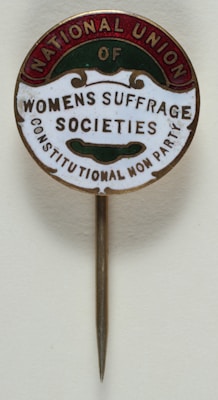A vintage pin with text that reads 'National Union of Women's Suffrage Societies, Constitutional Non Party'. The pin has a circular design with a metallic needle attached for wearing. The text is arranged within colored segments at the edges.