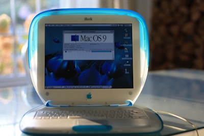 A vintage iBook laptop featuring a translucent blue and white exterior sits on a reflective surface, displaying the Mac OS 9 screen. Natural light filters through a window in the background, casting a warm glow.