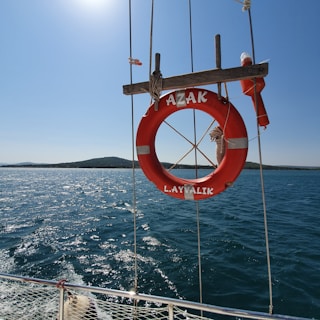 A bright red lifebuoy with white accents is hanging on the side of a boat, with a calm blue sea and a distant landmass under a clear sky in the background. The lifebuoy is secured by ropes and has the words 'AZAK' and 'L. AYVALIK' inscribed on it.