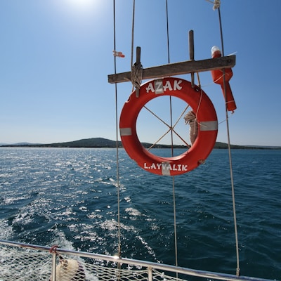 A bright red lifebuoy with white accents is hanging on the side of a boat, with a calm blue sea and a distant landmass under a clear sky in the background. The lifebuoy is secured by ropes and has the words 'AZAK' and 'L. AYVALIK' inscribed on it.