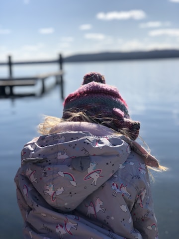 A child wearing a colorful knitted hat and a jacket featuring unicorns and rainbows stands with their back to the camera, looking out over a calm lake. The scene is serene with a clear blue sky and a wooden dock extending into the water.