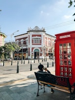A historic building labeled 'MARBA' is situated at a street corner. The architecture features a mix of red and white tones with decorative elements. In the foreground, a person sits on a bench beside a red, British-style phone booth labeled 'CHARGER BOX'. The area is paved and dotted with small trees in planters and black bollards.