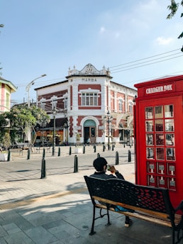 A historic building labeled 'MARBA' is situated at a street corner. The architecture features a mix of red and white tones with decorative elements. In the foreground, a person sits on a bench beside a red, British-style phone booth labeled 'CHARGER BOX'. The area is paved and dotted with small trees in planters and black bollards.