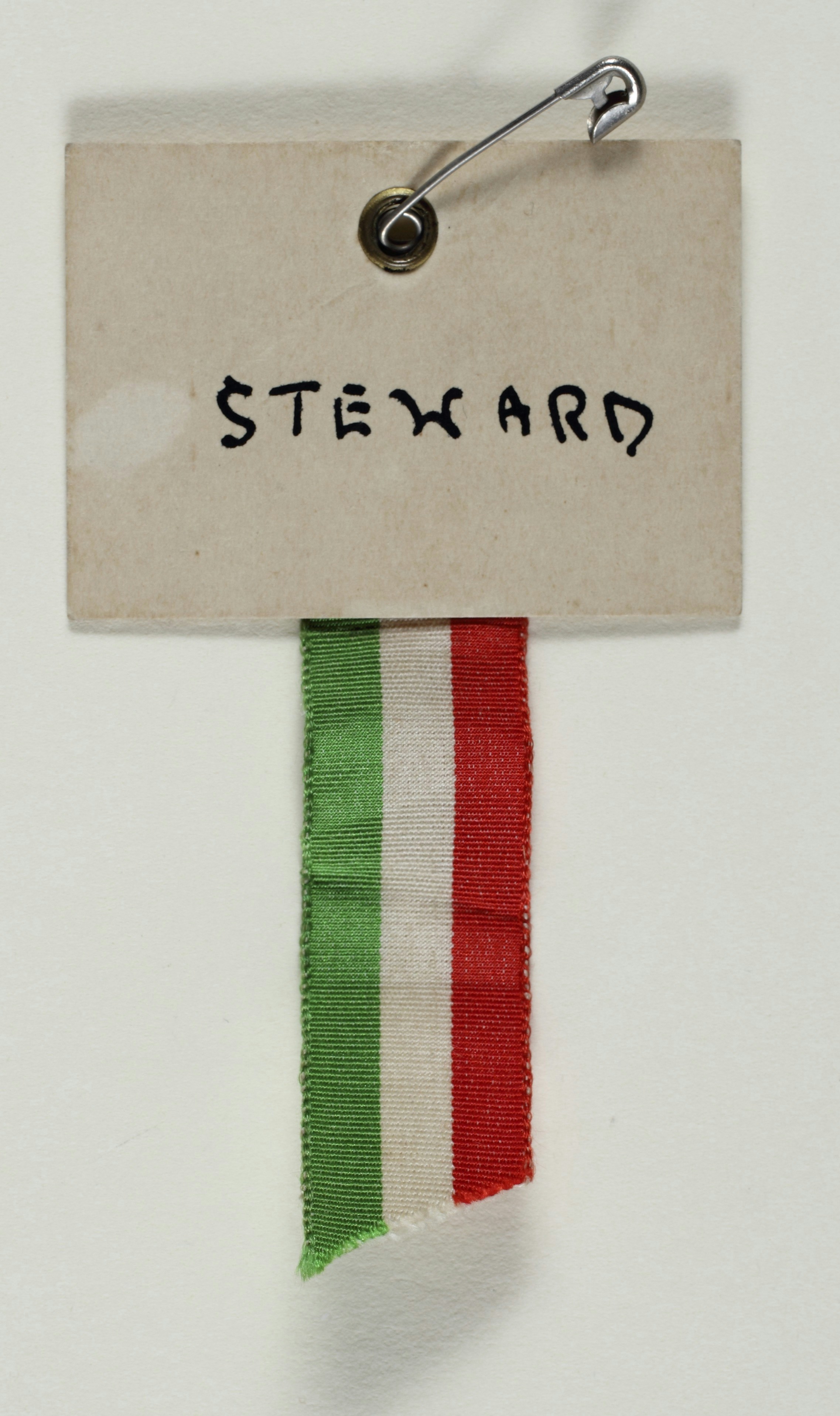 Suffrage Campaigning: National Union of Women's Suffrage Societies 1909-1928<br>Badge, cardboard, metal, textile, rectangle of cream cardboard, possibly produced by the National Union of Women's Suffrage Societies, manuscript inscription: 'Steward' in black ink, hole at top of cardboard for safety-pin to go through, reinforced, length of cotton, rep weave ribbon with red, white and green stripe design, attached to the back bottom edge of the cardboard, bottom of ribbon cut diagonally. TWL.2004.596.1