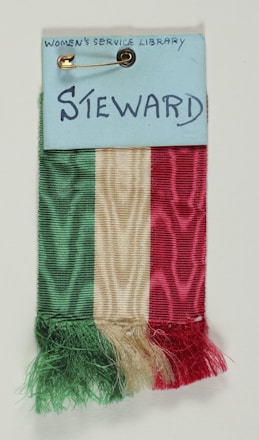 A ribbon composed of three vertical sections colored green, white, and red hangs from a card labeled 'Steward' with 'Women's Service Library' written at the top. The card is secured with a gold safety pin.