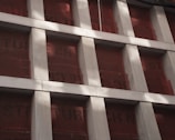 A partially constructed building facade with vertical concrete columns and red protective sheets behind them. The columns create a grid pattern, and text on the sheets is partially visible. Shadows from nearby structures or features fall across the surface.