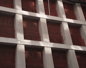 A partially constructed building facade with vertical concrete columns and red protective sheets behind them. The columns create a grid pattern, and text on the sheets is partially visible. Shadows from nearby structures or features fall across the surface.