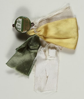 A vintage badge displays a green, circular button with the text 'Votes for Women' and an additional unreadable inscription beside three ribbons in green, yellow, and white.