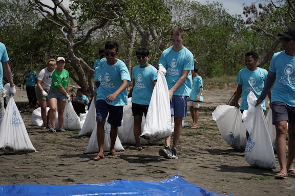 A group of people wearing blue T-shirts participates in a beach cleanup event. They are carrying large white bags filled with collected trash. The background consists of trees and a sandy beach, indicating an outdoor setting.