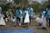A group of people wearing blue T-shirts participates in a beach cleanup event. They are carrying large white bags filled with collected trash. The background consists of trees and a sandy beach, indicating an outdoor setting.