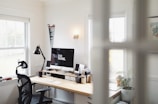 A bright and minimalist home office with a large window letting in natural light. The focal point is a computer with 'Do More' displayed on the screen, accompanied by various desk accessories like a lamp, potted plants, notebooks, and an office chair.