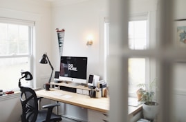 A bright and minimalist home office with a large window letting in natural light. The focal point is a computer with 'Do More' displayed on the screen, accompanied by various desk accessories like a lamp, potted plants, notebooks, and an office chair.